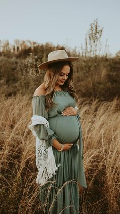 a pregnant woman wearing a green dress and hat standing in tall grass with her hands on her belly