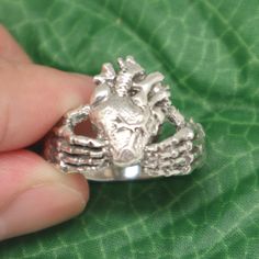 "Unique Claddagh Ring. It is made with a skull hand holding a real human heart. Cool! The Claddagh ring can be traced back to the ancient village of Claddagh. These rings were originally worn here as a sign of marital commitment and love. This ring takes the form of skeleton hands, grasping a hand carved anatomical heart and it is ideally adorned with the appearance of a silver crown, to give off an air of a modern Claddagh. This piece of art, is a perfect blend of ancient tradition in a modern Symbolic Hand-cast Skull Ring Gift, Real Human Heart, Hands Grasping, Heart Skull, Goth Ring, Halloween Ring, Ancient Village, Claddagh Ring, Skull Hand