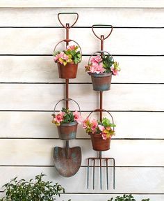 three pots with pink flowers are hanging on a wall next to a garden rake and flower pot holder