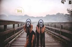 two people with masks on their faces sitting on a wooden bridge in front of the camera