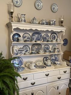 a white china cabinet with blue and white plates on it's shelves next to a potted plant