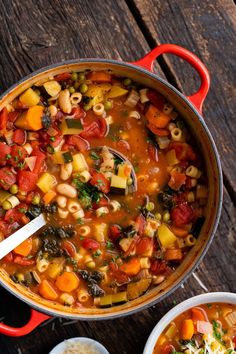 a pot filled with pasta and vegetables on top of a wooden table next to a spoon