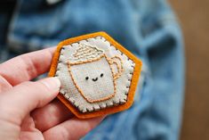 a hand holding a small embroidered badge with a coffee cup on it