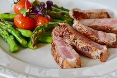 a white plate topped with meat and asparagus next to tomatoes, broccoli and cherry tomatoes