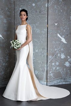 a woman standing in front of a wall wearing a white dress and holding a bouquet