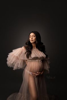 a pregnant woman in a sheer dress poses with her hands on her hips and arms behind her back