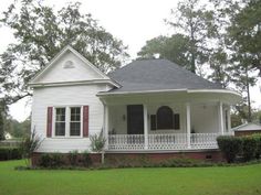 a white house with red shutters on the front porch and covered in grass, surrounded by trees