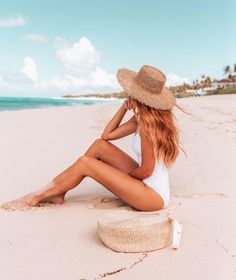 a woman sitting on the beach wearing a straw hat and white swimsuit with her legs crossed