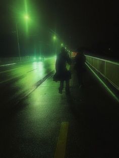 two people standing on the side of a road at night with green lights in the background