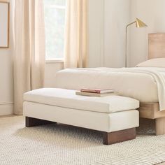 a white bed sitting next to a window with curtains on the windowsill and a bench in front of it