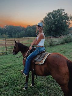 a woman riding on the back of a brown horse in a green field at sunset