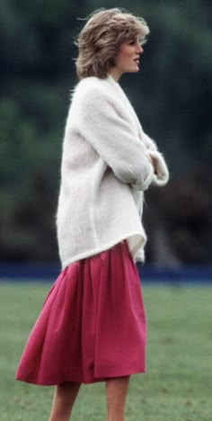 a woman in a red skirt and white sweater is standing on the grass with her hands behind her back