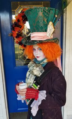 a woman with red hair wearing a green top hat and holding a cup in front of a blue door