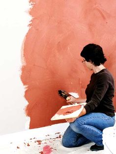 a woman is sitting on the floor with her paintbrush in hand and she is painting an orange wall