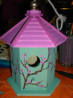 a birdhouse with pink flowers painted on the side and a purple roof is sitting on a table