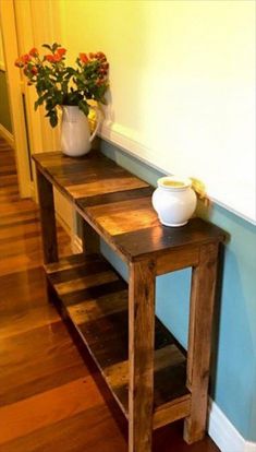 a wooden table sitting next to a white vase with flowers in it on top of a hard wood floor