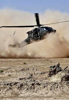 a helicopter is flying low to the ground with sand blowing around it and soldiers on the ground