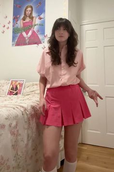 a woman in pink shirt and red skirt standing next to a bed with posters on the wall