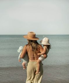 a woman walking on the beach with her child and baby in her arms while wearing a hat