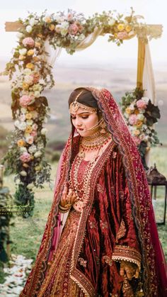 a woman in a red and gold wedding outfit standing next to a floral arch with flowers on it