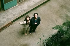 two people standing next to each other in front of a building with green doors and plants