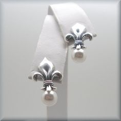 "Sterling silver fleur de lis stud earrings are accented with a glowing white Swarovski elements half-drilled pearl. Classic, beautiful and unusual. This symbol has traditionally been used to represent French royalty, and in that sense it is said to signify perfection, light, and life. Fleur de lis translates to \"flower of the lily\" in English. The earrings are about 7/8\" long (1.8 cm) from the top of the fleur de lis to the bottom of the pearl. Setting, earnuts and earring components are all Classic White Flower Earrings For Anniversary, White Classic Flower Earrings For Anniversary, Classic White Flower Earrings For Pierced Ears, White Classic Flower Earrings For Pierced Ears, Formal White Sterling Silver Flower Earrings, Classic Flower Earrings, Classic White Flower Earrings, White Classic Flower Earrings, Elegant Silver Clip-on Flower Earrings