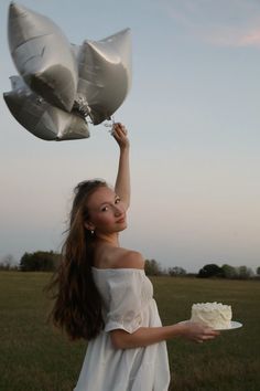 a woman in a white dress holding a cake with silver balloons on top of her head