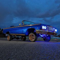 an old blue car is parked on the street at night with its hood up and lights on