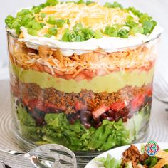 a layered salad in a glass dish on top of a white plate next to a fork and spoon