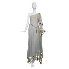 Stunning extremely rare Thierry Mugler Goddess Gown from one of his earliest Collections ca.1979. A masterful embodiment of ethereal elegance and avant-garde artistry. The dress cascades gracefully, draping effortlessly over the body. It can be worn either symmetrical or asymmetrical over one shoulder, the silhouette is both a nod to classical Grecian styles and a bold statement of modern femininity, capturing the duality of strength and delicacy that defines Mugler's aesthetic. Very soft, muted grey/champagne jersey cotton blend fabric. Highlight is the exquisite cutwork that adorns the hem and sleeves. These intricate patterns, meticulously edged in metallic gold thread, create a delicate interplay of light and shadow, transforming the fabric into a shimmering tapestry of opulence. Each