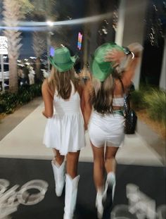 two women in white dresses and cowboy hats walking down the street together, both wearing green hats