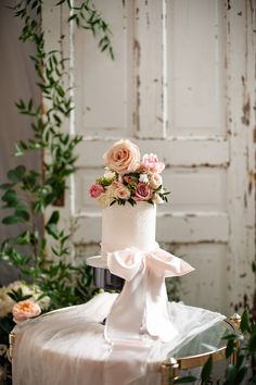 a white wedding cake with pink flowers on top sitting on a table in front of an old door