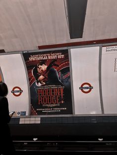 a woman standing in front of a movie poster at the subway station while looking at her cell phone