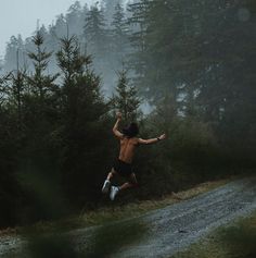 a shirtless man running down a dirt road in front of pine trees on a foggy day