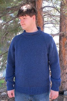 a man standing in front of some trees wearing a blue knitted sweater and jeans