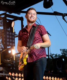 a man standing on top of a stage with a guitar in his hand and smiling at the camera