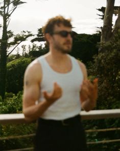 a man in white tank top and black shorts standing next to a fence with trees behind him