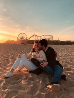 a man and woman are sitting on the sand at the beach with an amusement park in the background