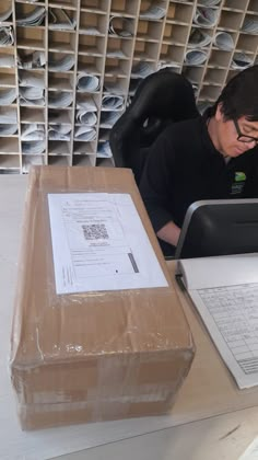 a man sitting at a desk working on a laptop computer next to a cardboard box