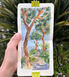 a person holding up a drawing in front of a tree and bushes with green clips