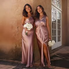 two beautiful women standing next to each other in front of a wall holding bouquets