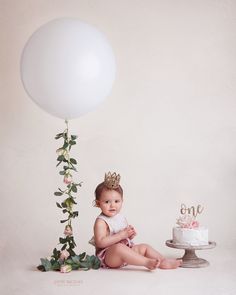 a baby sitting next to a cake and balloon