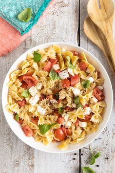 a white bowl filled with pasta and vegetables on top of a wooden table next to a spoon