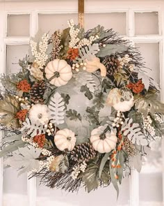 a wreath with white pumpkins, greenery and pine cones hanging in front of a window