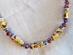 18" cloisonne necklace with gold and blue long round bead and orange crystal.  Lobster claw closure is 18 karat gold plate.  Accompanying pierced pendant earrings have a long blue and gold cloisonne bead with small gold beads suspended on an 18 karat gold plated stud post and back. This adds a quiet touch of sophistication to a simple blouse or tee.  Lampwork beads are hand made by skilled artisans melting and forming glass in a flame. Cloisonné beads are created when enamel decoration is applie Gold Beaded Necklaces As Costume Jewelry Gifts, Costume Jewelry With Gold Beads For Gift, Traditional Single Strand Gold Jewelry, Traditional Gold Single Strand Jewelry, Gold Costume Jewelry Necklace With Colorful Beads, Gold Single Strand Costume Jewelry, Gold Pendant Jewelry With Colorful Beads, Spiritual Gold Necklace With Decorative Details, Ornate Gold Beaded Jewelry