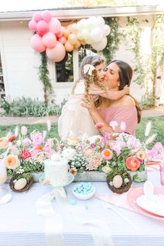 two women sitting at a table with flowers and balloons in the air, one kissing the other's cheek