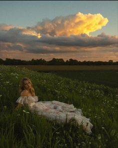 a woman sitting on top of a lush green field under a cloudy sky in a white dress