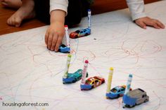 two children playing with cars and pencils on the floor in front of a large sheet of paper