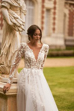 a woman standing next to a statue wearing a wedding dress with long sleeves and flowers on it