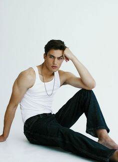 a young man sitting on the ground wearing jeans and a white tank top with his hands behind his head